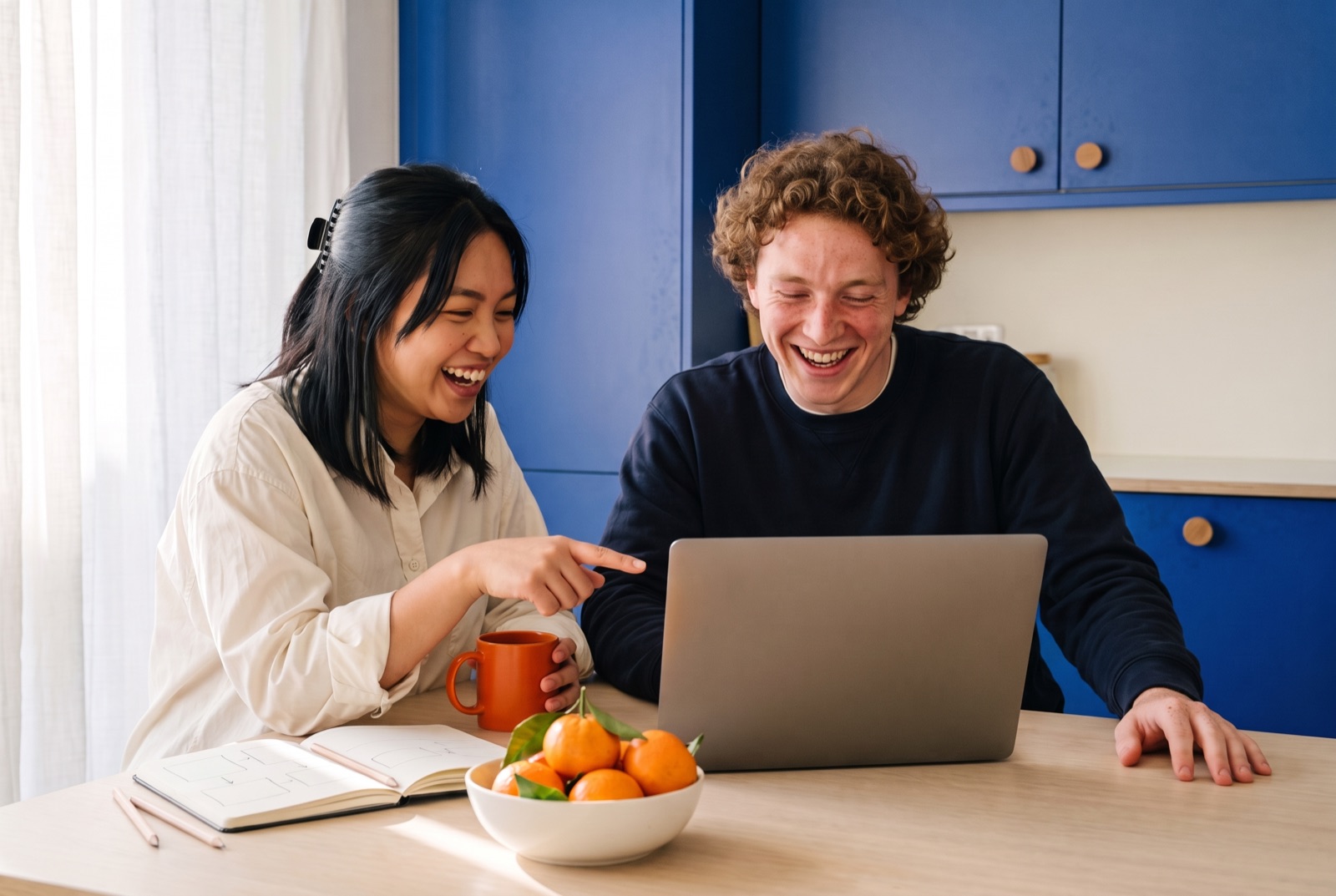 Two friends laughing at a laptop at a kitchen table, a notebook and a bowl of tangerines beside them.