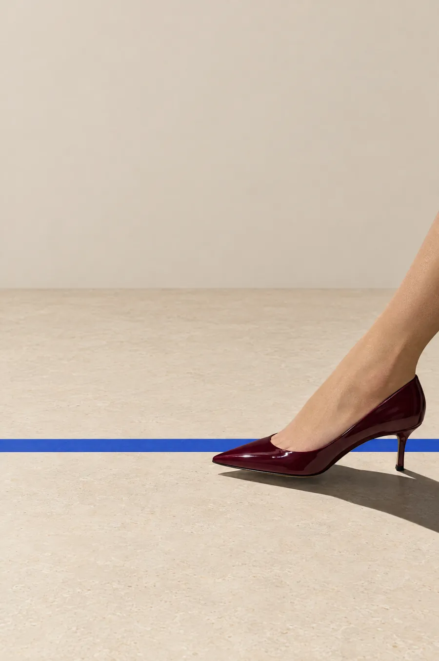 A woman’s oxblood patent heel stepping over a strip of blue tape on a pale stone floor.