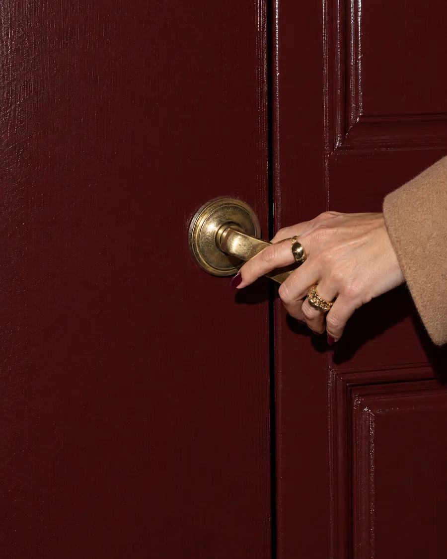 A woman’s hand with gold rings pressing down the brass handle of an oxblood front door.